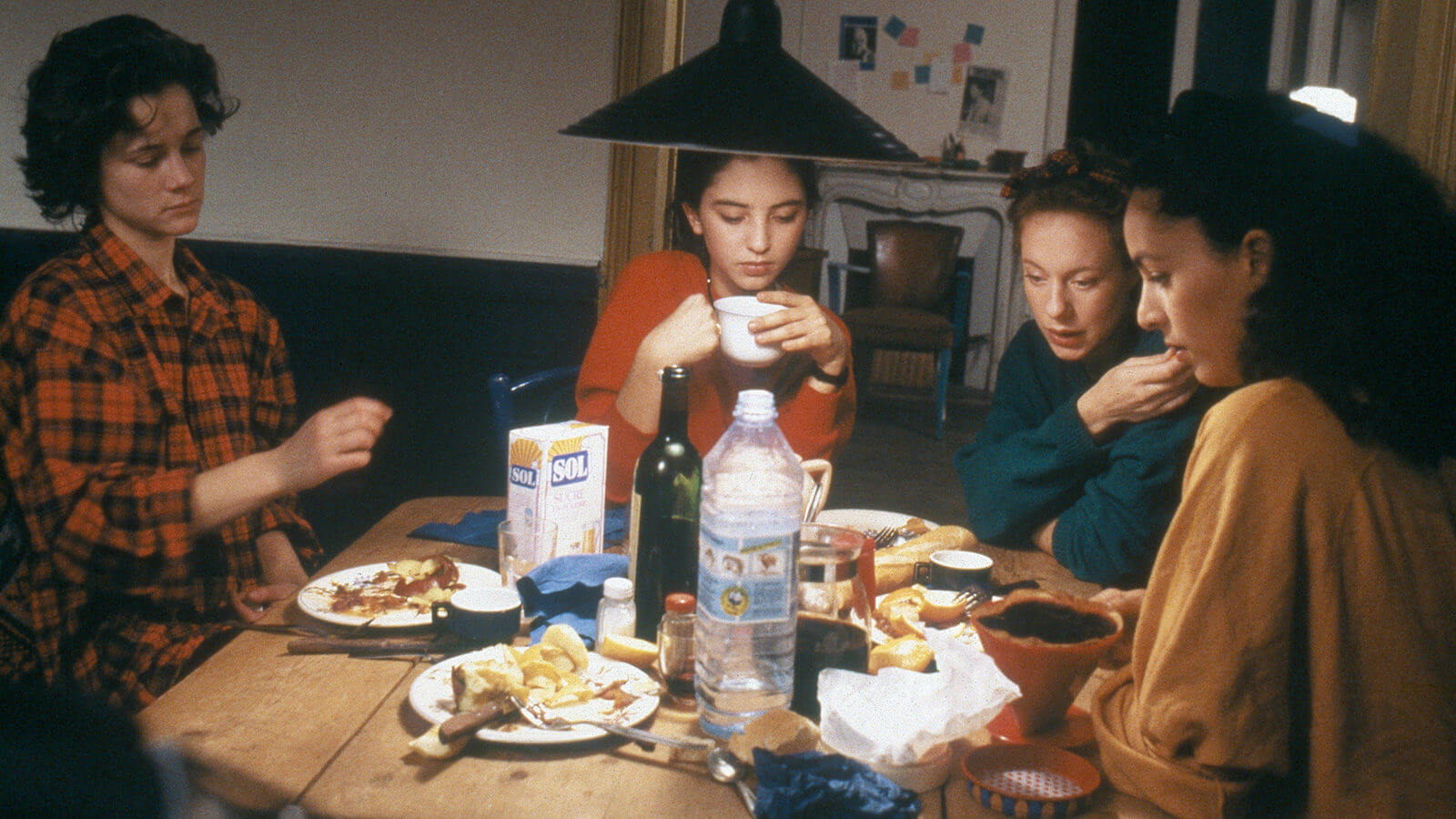 a still from the film Gang of Four depicting the four main characters dressed in flannels and jumpers sitting around a kitchen table cluttered with beverage bottles and half-eaten plates of food.